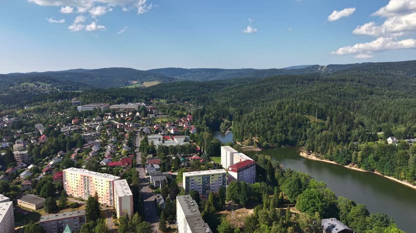 Aerial over town nestled beside winding river, bordered by dense pine forest and rolling hills. Visible are multi-story apartment blocks, mix of red and pale rooftops, tree lined boug