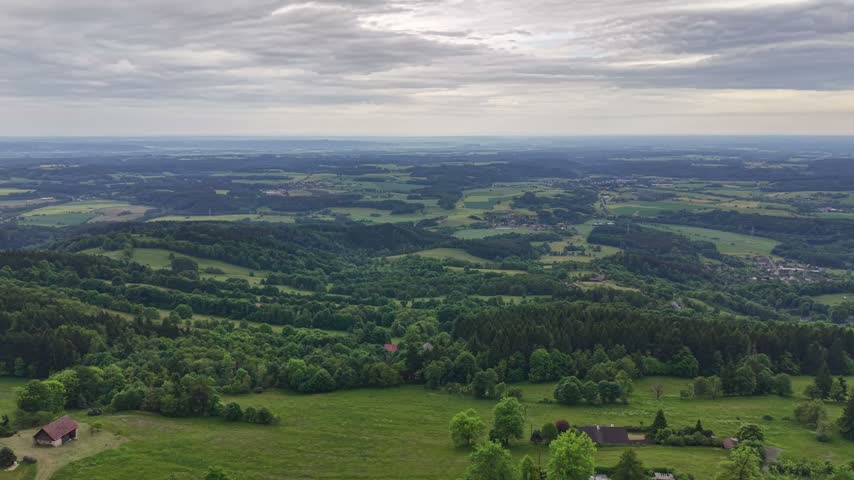 Aerial panorama over rolling countryside with patchwork fields, forested hills, and scattered farmhouses beneath expansive, overcast sky. Subtle wind movement stirs trees and grass as view glid