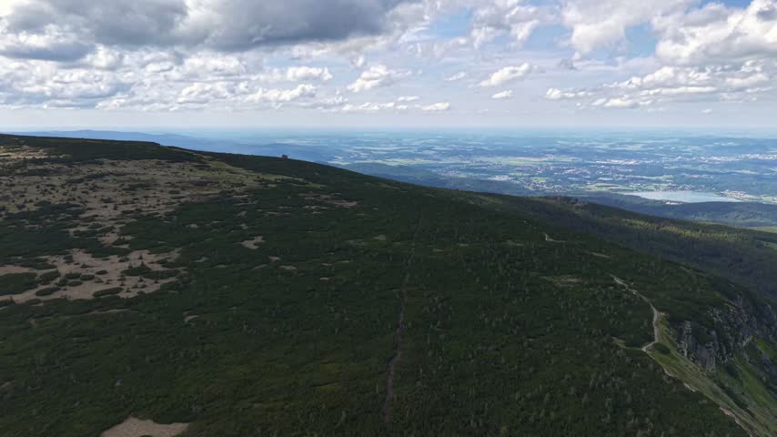 View from hillside shows winding dirt trail through low shrubs and evergreen patches, leading eye toward distant valley beneath shifting cloud cover and light shadows, wide panoramic calm.