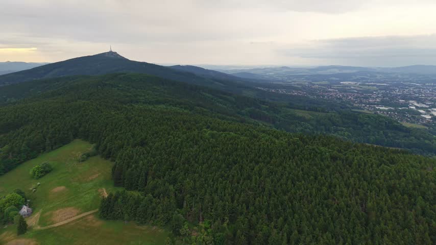 Aerial motion shows lush green forested hills leading to distant town in broad valley under soft, diffused daylight. Rolling ridges, mountain summit with small structure at peak, and open