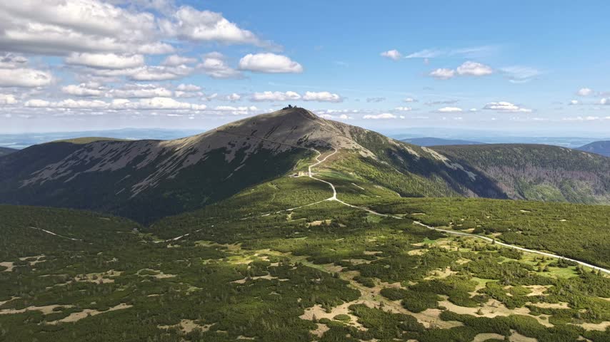 Winding dirt trail climbs sunlit mountain ridge under bright blue sky with scattered white clouds. Lush green shrubs cover lower slopes, giving way to rocky, barren summit. Panoramic landscape