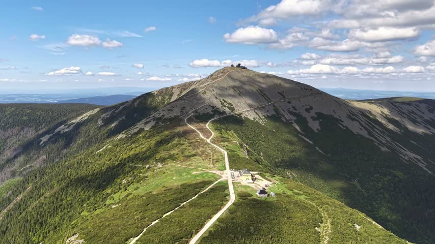 Aerial mountain landscape with winding dirt trail climbing toward summit and small buildings at peak. Bright blue sky with clouds, green vegetation, and rocky slopes view for travel and hiking.