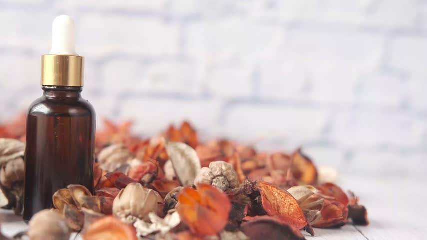 Close up video of essential oil dropper bottle and face cream jar placed on wooden table with dried potpourri leaves, featuring smooth panning camera motion for cosmetic product display.
