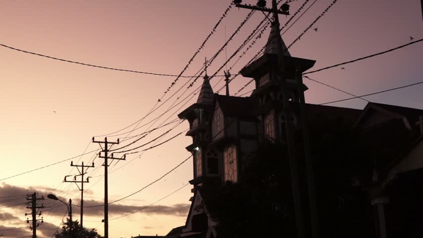 Silhouettes of a church building and electric poles under a dramatic sunset sky with birds flying and perching on wires