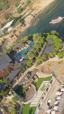 Aerial view of a landscaped resort with swimming pools, tiled roofs, rocky shoreline, and turquoise coastal water.