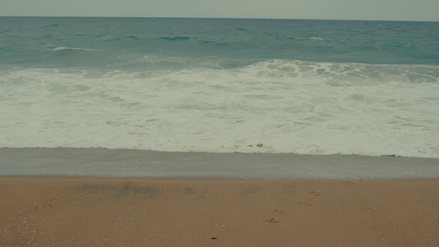Rolling ocean waves approach the sandy Newport Beach shoreline under a gray and cloudy coastal sky.