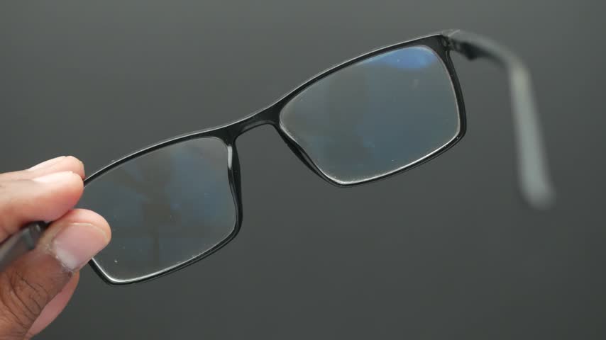 Person holding black frame eyeglasses and spraying clear lens cleaning solution onto surface of glasses to sanitize and remove smudges for clear vision in home setting.