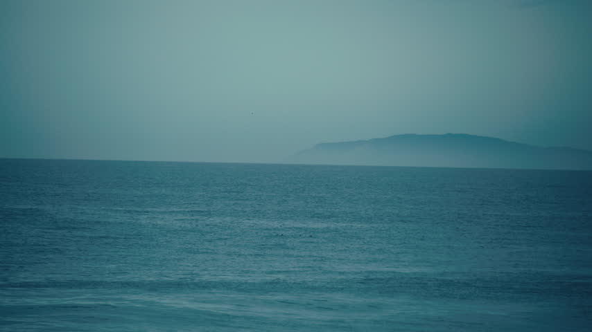 A distant island rises above the calm blue ocean horizon beneath a hazy sky off the California coast