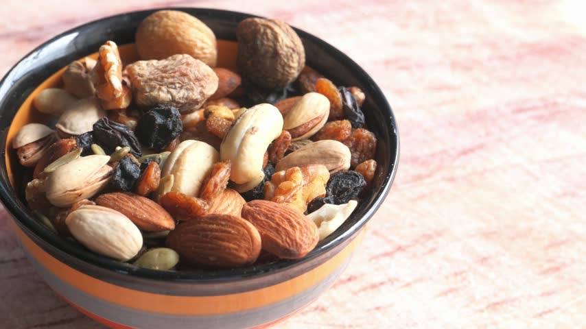 Top view of a small bowl containing a variety of nuts including almonds cashews pistachios and dried raisins with figs placed on a rustic light wooden table surface in soft natural lighting.