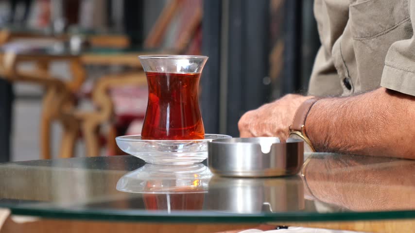 Glass cup filled with hot red turkish tea on glass table outside. Senior man with watch sitting near ashtray with cigarette at outdoor cafe. Close up shot of relaxing moment.