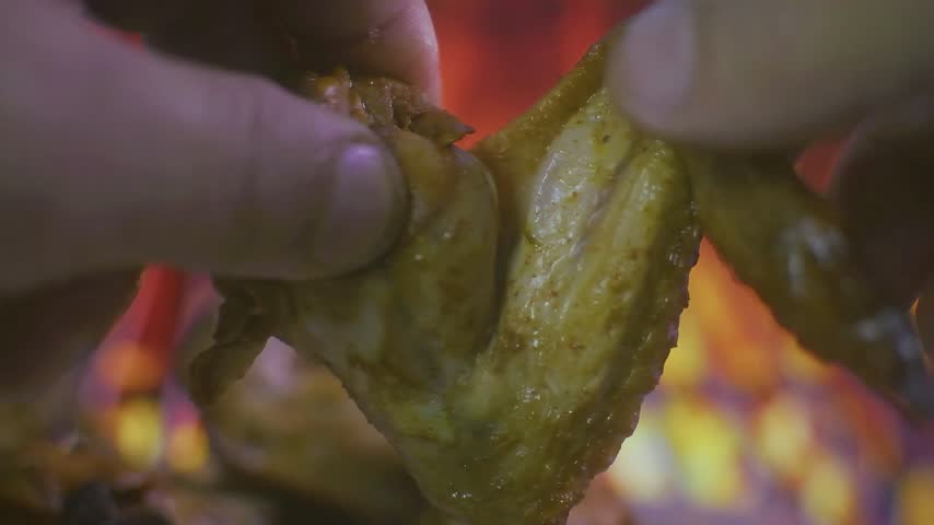 Hands are pulling apart a cooked chicken wing, revealing the juicy meat inside. The background is blurred with orange and yellow flames.