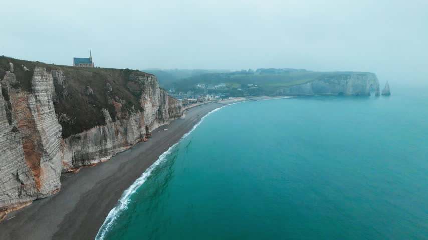 Rain and coastal haze soften distant cliffs at Etretat, Normandy, France. Drone footage of chalk headland above pebble beach and bay.