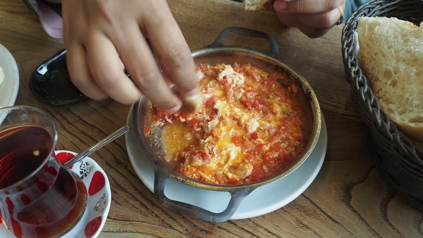 Person dipping fresh bread into traditional Turkish menemen eggs with tomato and peppers served in hot copper skillet on wooden table, close up shot with tea on side.