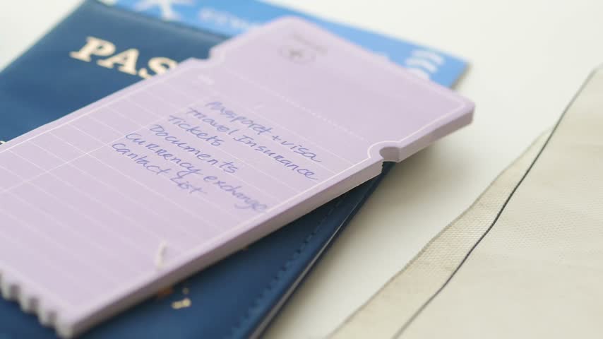 Handwritten travel checklist note resting on passport alongside miniature airplane stencil and compass on white surface. Close up overhead shot depicting organized trip planning.