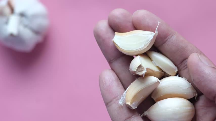 Close up shot showing hand holding bunch of peeled garlic cloves placed on palm against vibrant pink background during food preparation process for healthy cooking recipes.