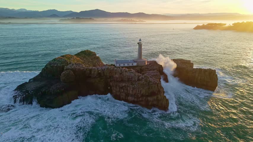Aerial view of the Mouro Island lighthouse situated on a rocky islet in the ocean, with crashing waves and golden sunset light in the background, Santander, Cantabria, Northern Spain.