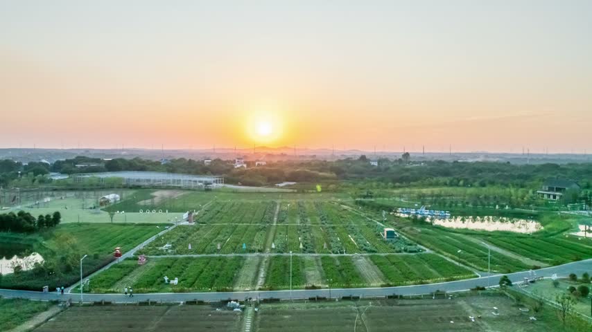 Sunset time lapse drone aerial view of green rural countryside with distant mountains
This is a beautiful sunset time lapse photo taken from a drone. The aerial view reveals expansive green fields and organized crop rows with shifting light and shadows under the warm twilight. Distant mountains are clearly outlined against the orange-red sky, while greenhouses, ponds, and paths form a peaceful yet dynamic rural scene.