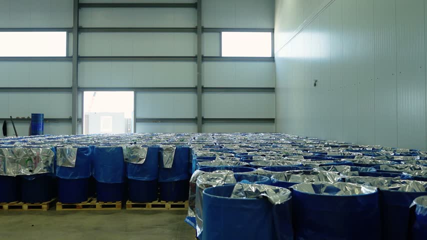 Large rows of blue plastic drums lined with silver foil sit on wooden pallets across a spacious industrial storage hall with a bright doorway at the back.