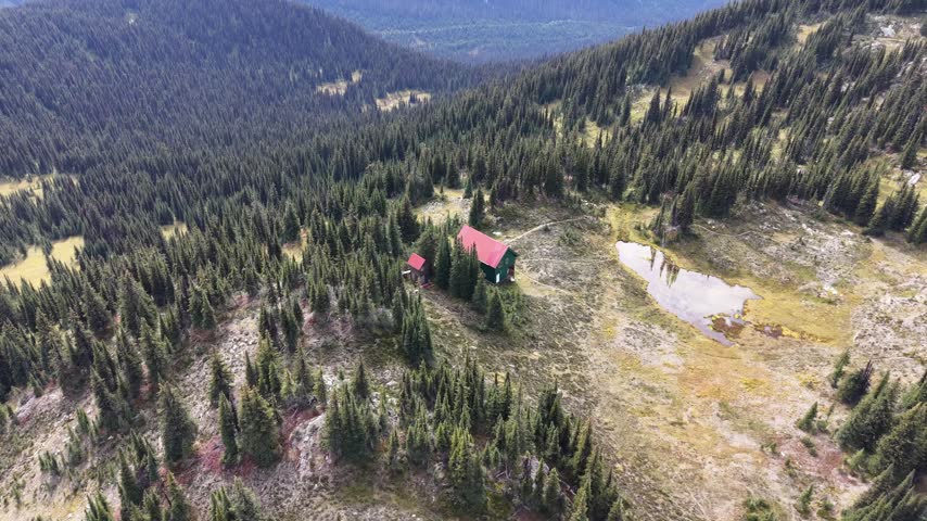 The camera circles a small red-roofed shelter in an alpine clearing on Trophy Mountain, Wells Gray Provincial Park, British Columbia, Canada, with nearby ponds, conifer forest and rugged peaks.