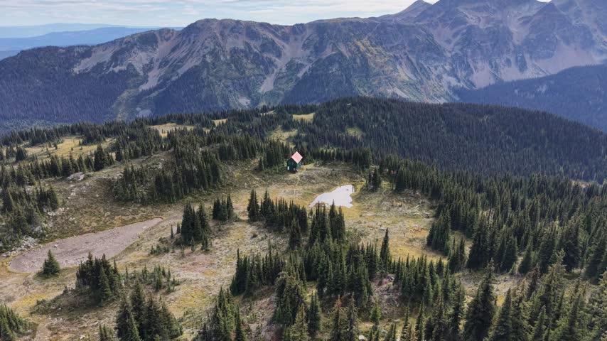 A drone reveals a small red-roofed mountain shelter beside alpine ponds on Trophy Mountain in Wells Gray Provincial Park, British Columbia, Canada, surrounded by open meadow, conifers and distant peaks.