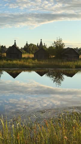 Wooden huts and church towers stand beyond a pond, with reeds along the water and clouds reflected on its surface in the evening.