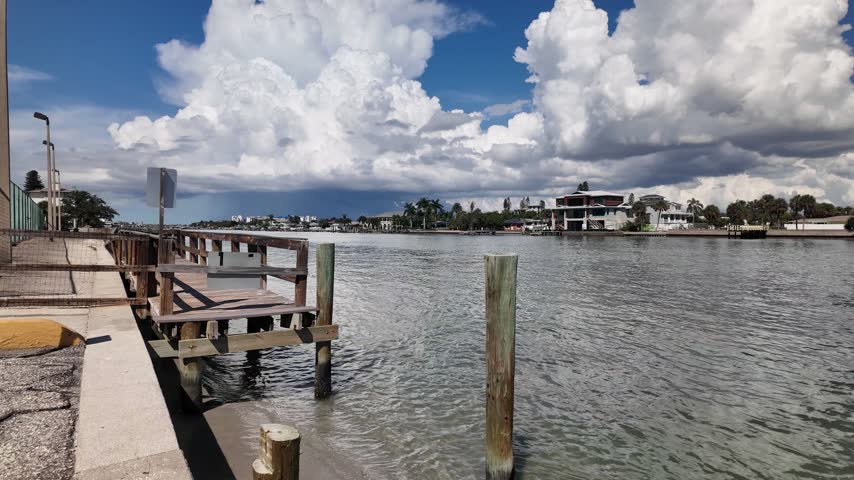 A wooden boardwalk runs beside a calm coastal canal in Treasure Island,FL as dramatic storm clouds gather overhead, with waterfront homes, docks and a small boat visible in the distance, conveying tension and anticipation.
