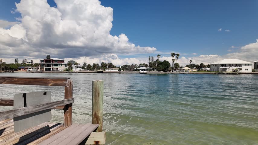 A wooden boardwalk runs beside a calm coastal canal in Treasure Island,FL as dramatic storm clouds gather overhead, with waterfront homes, docks and a small boat visible in the distance, conveying tension and anticipation.
