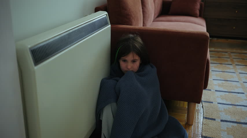 Freezing girl wrapped in a warm knitted blanket sitting on the floor between the sofa and electric radiator, trying to warm up in a cold apartment during winter.