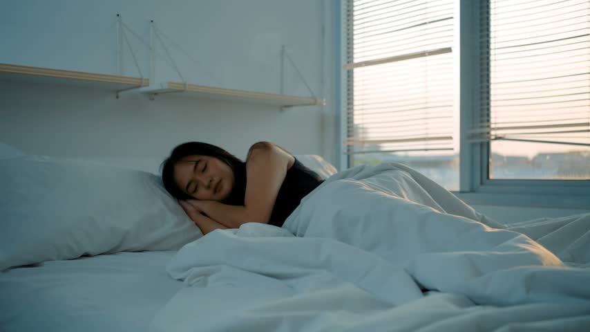 Beautiful young asian woman waking up in the morning and stretches in the bed radiated sunshine from the big window.
Happy young girl greets new day with warm sunlight. 4k video.