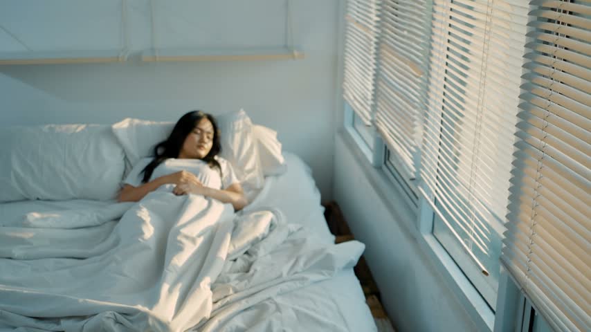 Beautiful young asian woman waking up in the morning and stretches in the bed radiated sunshine from the big window.
Happy young girl greets new day with warm sunlight. 4k video.