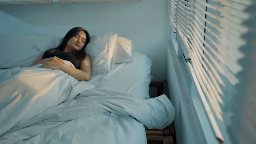 Beautiful young Asian woman stretching her arms and waking up in bed near a bright window. 4k video, slow motion.