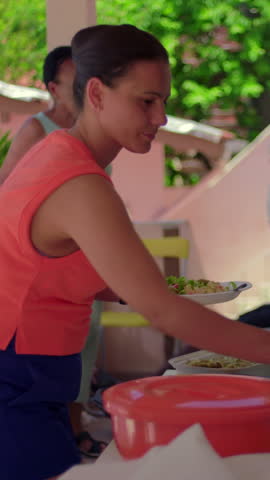 Young woman adds dressing from a squeeze bottle onto a fresh salad plate at a food buffet while preparing lunch during a summer family barbecue