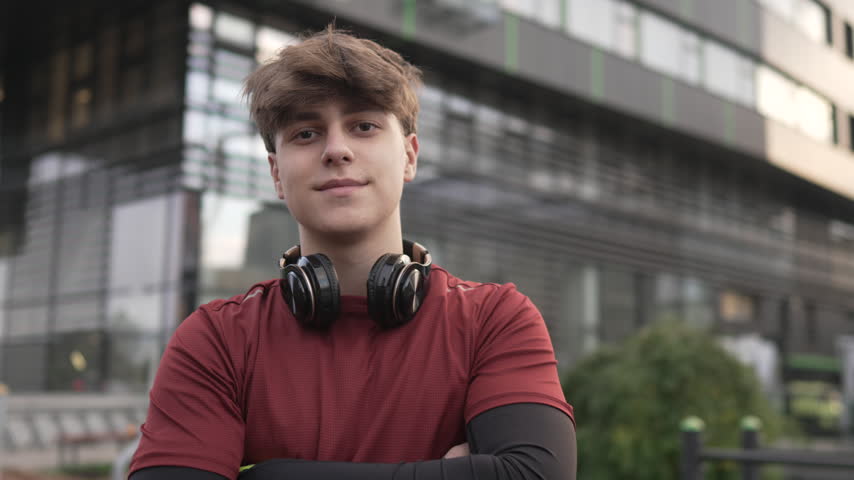 Portrait of a handsome young man in sportswear with headphones around his neck, standing with arms crossed and looking at the camera in a modern urban environment with a confident smile