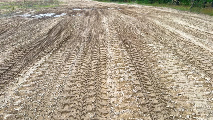 Parallel tire tracks cross a muddy gravel field with shallow puddles beside a forest, wet ground texture, vehicle imprints, rural terrain.