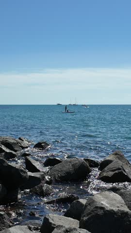 Vertical coastal view of Lido di Ostia near Rome, Italy. A person enjoys stand up paddleboarding on the blue Tyrrhenian Sea, with sailing yachts in the distance and large dark rocks along the shoreline. Gentle waves sparkle in the sunlight beneath a clear summer sky.