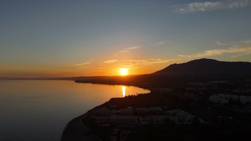 Aerial drone view of a dramatic sunset with the sun setting directly over the sea horizon, casting a silhouette over a curved coastline and mountain, near Bel Air, Estepona, Costa del Sol, Spain. Golden and orange light reflects across the calm water as darkness falls over the landscape below, marking the end of a clear day.