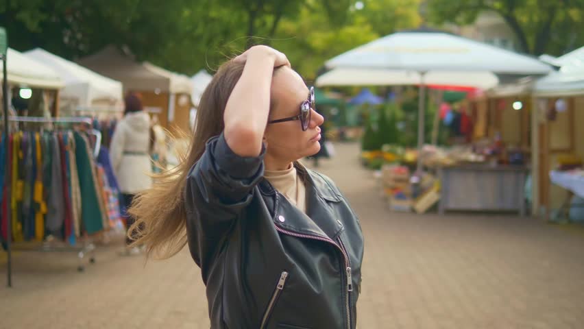 Woman in leather jacket walks through outdoor market. She wears sunglasses and runs a hand through her hair. Background features tents, clothing racks, and shoppers