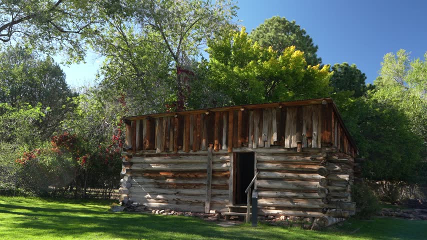 A rustic log cabin with a vertical wood upper section sits on a grassy lawn surrounded by trees under a clear blue sky.