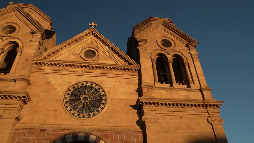 Low angle view of the Cathedral Basilica of St. Francis of Assisi in Santa Fe, New Mexico, featuring twin towers and a rose window illuminated by warm sunlight.