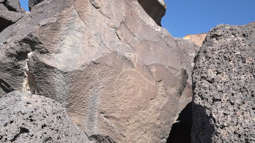 Close-up view of ancient rock carvings etched into the surface of massive, dark volcanic rocks under a clear blue sky.
