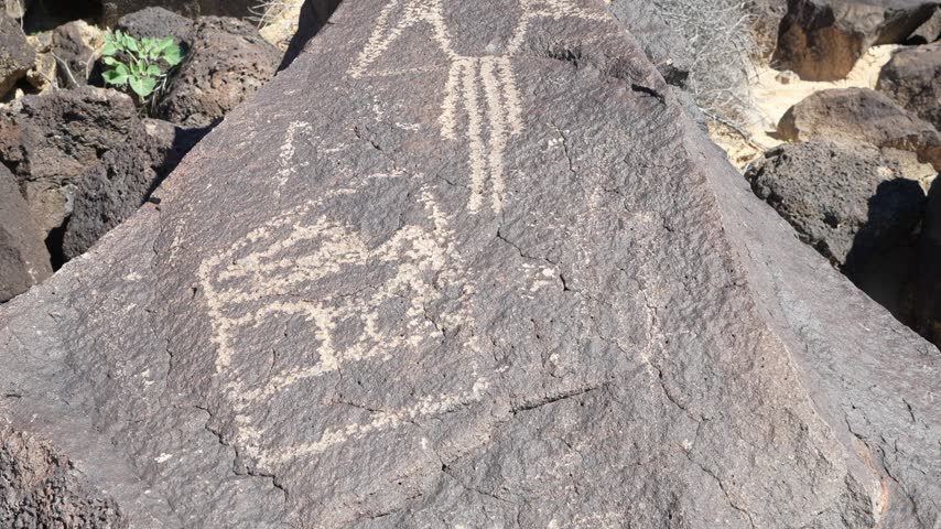 Close-up view of ancient rock carvings depicting abstract figures on a large boulder surrounded by desert terrain.