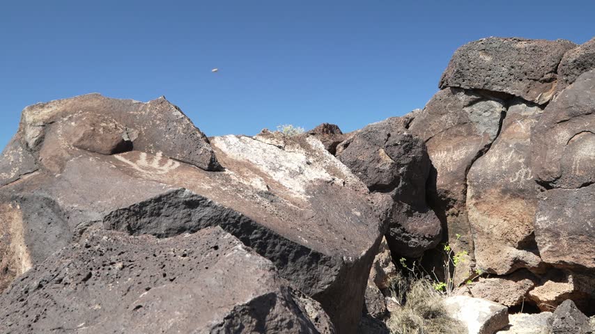 Sunlight illuminates a rugged landscape of dark, porous volcanic rocks featuring ancient petroglyphs, including a distinct spiral design, under a clear blue sky.