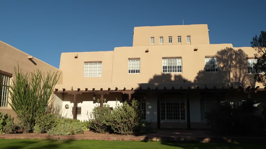 A large tan adobe building with wooden porch beams and desert plants is shown under a clear blue sky.