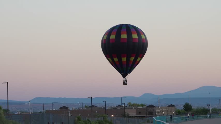 A large hot air balloon with a black and multicolored checkered pattern floats high above a city skyline and distant hills during twilight.