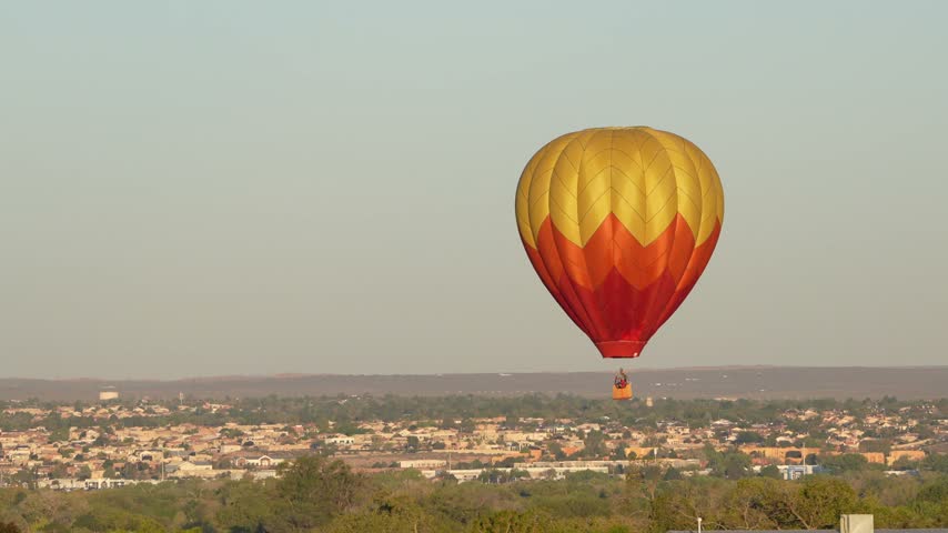 A yellow and red hot air balloon drifts through the sky above a sprawling city landscape and distant hills.