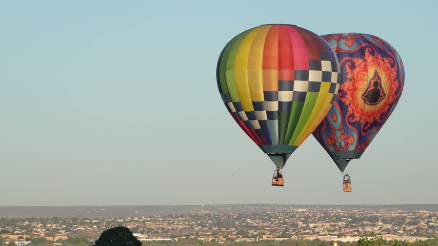 Two vibrant hot air balloons, one with a checkered pattern and one with an ornate design, drift above a sprawling city landscape.