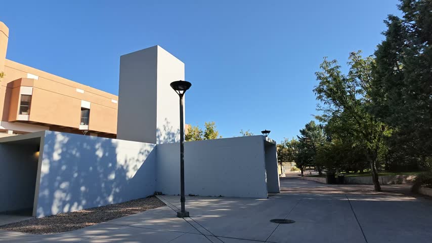 A minimalist concrete wall and tower structure stands on a paved campus walkway under a clear blue sky.
