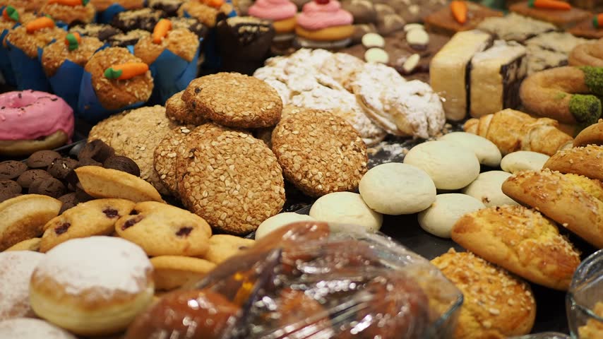 Close up cinematic footage captures wide variety of artisanal bakery products including cookies donuts cupcakes and muffins neatly presented on display for sale in cafe environment.