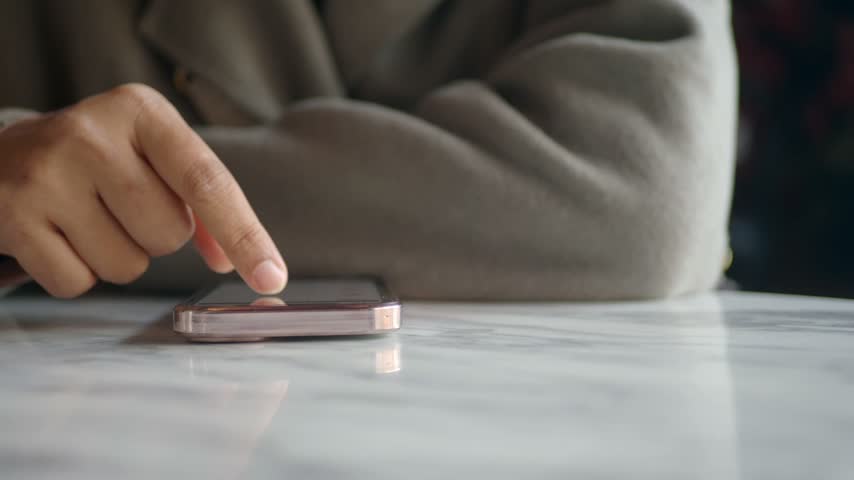 Close up shot of person hand scrolling and tapping on smartphone screen while resting on polished white marble surface in bright cafe environment.