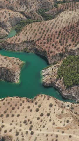 Aerial view of a winding turquoise reservoir surrounded by dry orchards, rugged cliffs, and forested slopes.